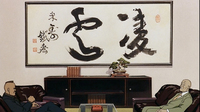 Black calligraphy in a frame on the wall of an office dominates the film frame, where two men sit conversing. One man sits in a leather chair in the left bottom corner of the frame facing a man sitting in the corner bottom right of the frame, also facing back at him. A coffee table is directly between them, with a bookshelf behind it and them in the midground. A few books and a bonzai tree are on top of it The massive calligraphy reads "ryoun" (凌雲), and means "above this mortal world."