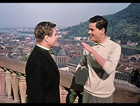 Walter talking to the chaplain while standing on a bridge above the city, holding his trumpet case under his arm.