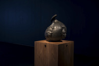 Still photo: Close-up of a black glass sculpture in the shape of a teardrop or conch shell with a human face protruding from the front and right-hand side. The sculpture is sitting on a medium-brown wooden pedestal.