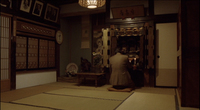 A person sits alone in a room, in front of an altar. Calligraphy can be seen on signs and scrolls throughout the room.