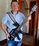 A man holds a gun-shaped guitar with a strap resembling a bandolier over his shoulder. His face is pixelated to protect his identity.