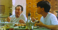 An older man and a teenage boy sit at a table eating in white t-shirts, in front of dozens of small wooden drawers in the background.