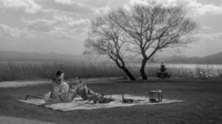 A black-and-white film still of a long shot, showing two small figures picnicking beneath a tree in an open field. The seated female figure holds a hand drum. The half-reclining male figure gazes at the female figure.