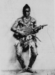 Black and white publicity photo from 1931 of performer Habib Benglia dancing. His costume incorporates body paint, shells, feathers, and beads. He is playing an Algerian stringed instrument while bending his knees in a deep plié.