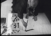 A man paints calligraphy on white paper, in black and white cinematography.
