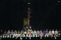 Figure 5.3: A color photograph of a theater stage with a stepladder, kettles, and buckets upstage, and confetti falling on nineteen performers sitting on a line of chairs downstage.