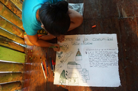 A young indigenous girl painting a scene of her community of origin, with a river, trees with fruits, birds, a few houses, a central ceremonial structure, a soccer field, and a text describing daily activities.