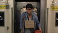 A man in a subway car wears a piece of cardboard around his neck with black calligraphy written on it. Signs on the wall have green calligraphy printed on them.