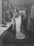 Photograph showing Blanche Bates in a dressing room looking at herself in a mirror while wearing a kimono and holding a fan