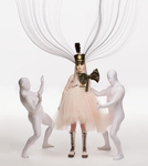 Poppy, wearing white combat boots, a beige dress with oversized black bow, and a black band-­leader hat, stares into the camera, while two men in white morphsuits flank her.