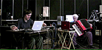 Color photo, two people are seated at tables in front of laptops, surrounded by audio equipment, including an accordion and audio interfaces.