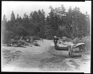 A black and white photograph of a group of people on the shore of a lake. A group of men relax on the sand as a woman sits in a canoe and a man points a gun across the lake.