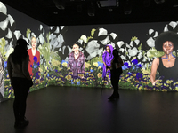 Photo: A large digital installation. There are three walls with videos being projected onto them that give the visual effect of layers. The background layer is a contrast of black with large white petals from a cotton plant. In the foreground are purple-and-yellow poppies and yellow roses. In the middle, there are four Black women of different ages, each wearing a different garment to represent the time period she represents. In front of the video projections are two viewers interacting with the artwork.