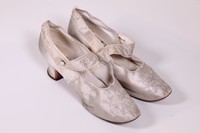 Pair of women's cream-colored satin wedding shoes, with modest heels, tone-on-tone floral embroidery, and a buttoned strap across the instep.