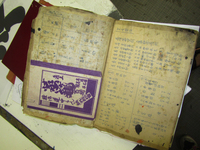 A photo of an old and yellowed book opened to pages showing charts and details of calligraphic characters. A purple and white pamphlet has been paper clipped to the interior of the left page.