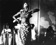 Photo of upright Afro-Puerto Rican dancer and scholar, left arm across her chest, right arm placed behind her waist, musicians behind her.
