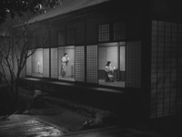 A long shot from a black-and-white film, showing two small female figures dressed in kimono, lighting candles in a Japanese-style room with three open doors. Each figure is framed by one of the open doors.