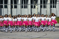 A large group of (mostly) young women dressed as schoolgirls with white shirts, bright pink skirts, white socks, and black shoes march in the middle of a street. They all have prosthetic pink lips that are partly open. A gray building is on the background