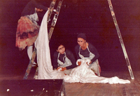 A maid dressed in blue and wearing a white apron is going up a folding ladder. Two other maids dressed in blue and wearing a white apron are on their knees, talking and holding a white dress.