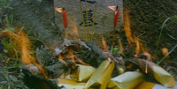 Image of objects burning in front of a tombstone, which has black calligraphy engraved into it.