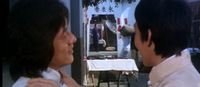 Two characters talk to each other; in the far background is some calligraphy over an entranceway.