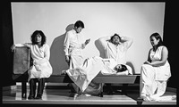 Black-and-white photograph. Five performers sit on a bench, dressed in white; the Māori character sits separately, guarding her suitcase.
