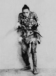 Black and white publicity photo from 1931 of performer Habib Benglia dancing. His costume incorporates body paint, shells, feathers, and beads. His hands are in front of him, as he is about to pounce.