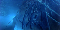 Worm's-eye view of a tombstone with calligraphy on it and dead vines around it. The scene is blue-tinted.