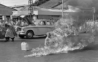 Self-­immolation of Thích Quảng Đức, South Vietnam, 1963. Thích Quảng Đức sits in meditative lotus position in the middle of the photograph, flames blasting to his right from his head and body. He is seated in the middle of two lines of flames. A white petrol cannister sits on the ground to the left. In the immediate background sits a 1950s-­style car with its hood up. Đức’s burning body is surround at a distance by monks with shaved heads and traditional Buddhist shawls. Behind them are other onlookers and city buildings of Saigon.