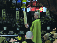 Fig. 17. A photograph of a temple man reading from a book held up. Other people are in the foreground and flowers and offerings are visible in the background.
