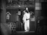 A man stands in a street, holding a fan up. Calligraphy can be seen on the shop curtains and lanterns.