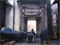 Film still showing a young woman lounge against a large stone while a man in a chair watches her.