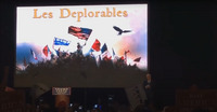 Donald Trump stands behind a podium. On the screen behind him there is an image of the barricade scene from the musical “Les Misérables” with a Trump banner, the American flag, and a bald eagle added. The words “Les Deplorables” appear at the top of the screen in yellow.
