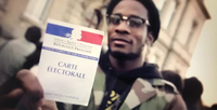 Still from a musical campaign video. A young Black man holds up his voting credential card.