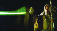 A color still from a film. It shows an old male figure with a long staff in his left hand, holding up an alms bowl in his right hand to absorb a laser-like greenish light.
