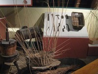 Photo of museum display showing an incomplete basket in the foreground, with tools and other baskets in the background.