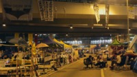 A still from the film “Yellowing” shows a protester’s village at night. Tents and tables are set up beneath an overpass. Few people are out, but on the right, a small group sits in folding chairs, office chairs, and on boxes