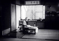 A man kneels holding a paper with a banner hung on the wall above him with black calligraphy, in black and white cinematography.