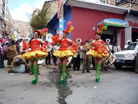 Three drag queens in heavy makeup, green platform boots, and red-and-yellow skirts, corsets, and top hats smile and wave in front of a brass band during a procession.
