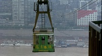 A cable car crosses over a river towards the city beyond. Yellow calligraphy is visible on the car itself.