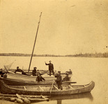 A black and white photograph of seven men standing or sitting in a small fleet of birch-bark caones by a dock on a lake.