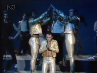 Figure 19. As Sha Na Na perform the teen tragedy song “Tell Laura I Love Her,” three singers in gold lamé suits stand at the center. Two form the roof of a chapel by raising their arms, while the third is on his knees singing beneath.