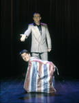 Robin Arthur in a rectangular plastic shopping bag in the colors of the British flag. He is kneeling into the bag and only his head is visible above the bag’s zipper. Standing behind Robin, John Rowley smiles at the audience. The rectangular shopping bag recalls the “black box” aesthetic exemplified by Rauschenberg’s Music Box.