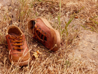 An outdoor close-­up of a pair of used Mekap shoes in the Qandil Mountains, Kurdistan. The shoes are amber-­brown with loosely tied laces, almost camouflaged against the brown earth beneath them.