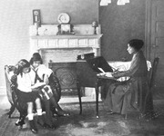 This sentimental photograph by an unknown photographer of a domestic scene depicts a mother and her two daughters in a well-appointed middle-class home complete with a lush area rug, carved marble fireplace, polished writing desk, and armchair. Both the mother and two daughters have straightened hair and wear carefully starched formal attire to indicate their respectability.