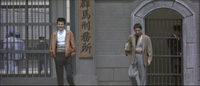A man leaves a prison gate, while another man stands nearby. A large calligraphy board can be seen in the background behind them.