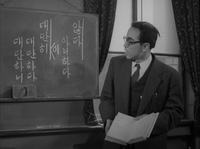 A professor holds open a book in front of a chalkboard with white calligraphy written on it, in black and white cinematography.