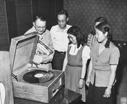 Donald Beckman Brown, chief of the Information Division of GHQ, stands with Japanese high school students. All are smiling and gazing at a turntable. Brown is holding a recording of the Broadway musical Oklahoma!