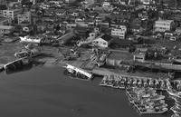 A neighborhood is visible along the top of the photograph, with a few damaged buildings and houses. Below is a partially destroyed dock and several capsized and grounded ships. To the bottom right are many more, smaller ships moored to the undamaged part of the dock.