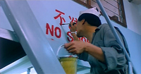 A low angle shot shows Mark on a ladder painting bilingual red characters that say "No Smoking" on a wall.